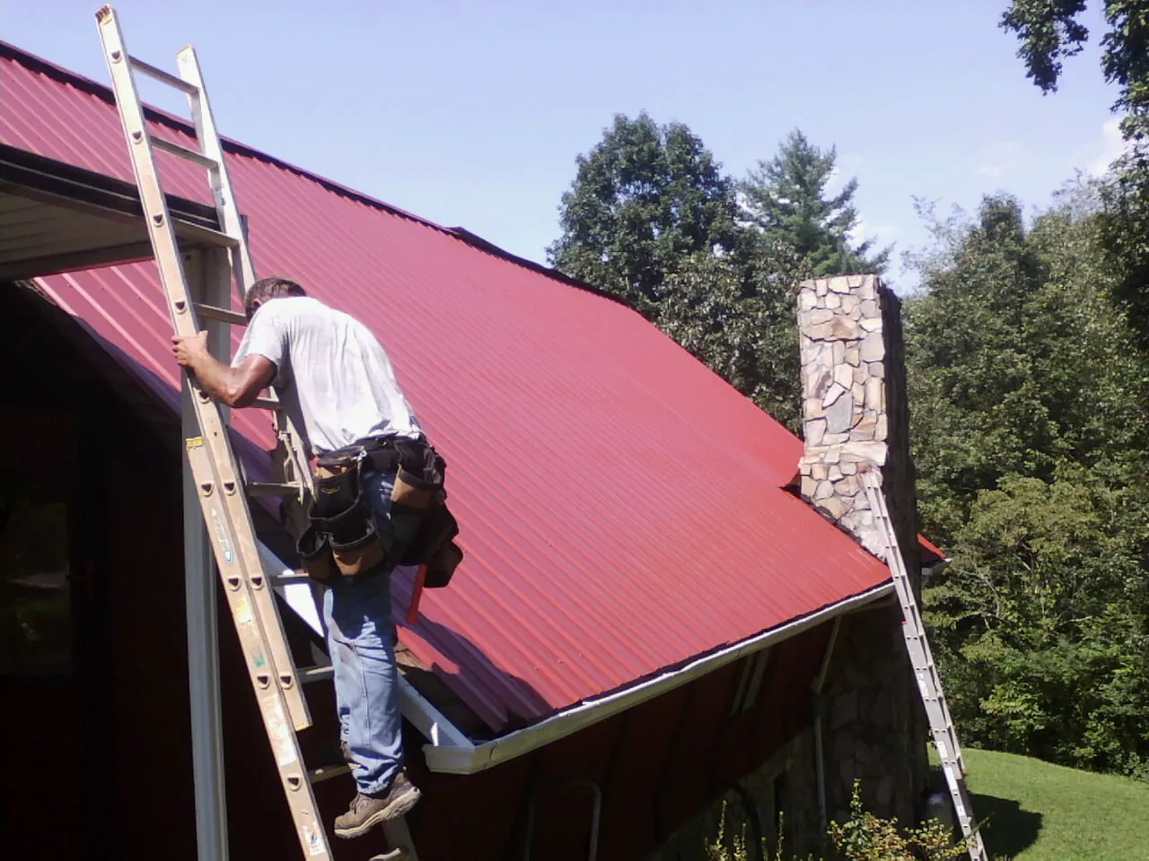 Roofer on a ladder beside a red metal roof and stone chimney — Soffit & Fascia Repair by Roofing Brothers in Glen Dale